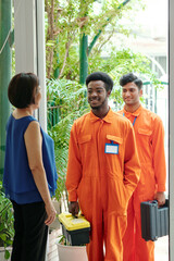 Mature Asian woman standing at house entrance and smiling to repair workers while greeting them