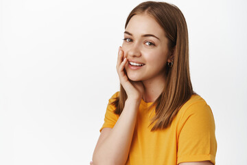 Portrait of beautiful teen girl, touch skin without acne, no make up, touch cheek gently and smiling tender at camera, standing in yellow t-shirt against white background