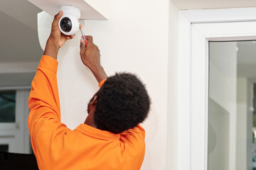 Rear view of black handyman in orange wear using screwdriver while fixing security camera on ceiling