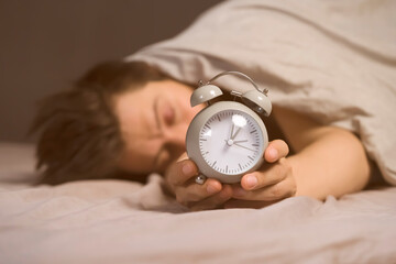 Hands of a young man from under the blankets hold a retro vintage alarm clock in gray. The person...