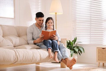 Father and daughter reading book on sofa at home - Powered by Adobe