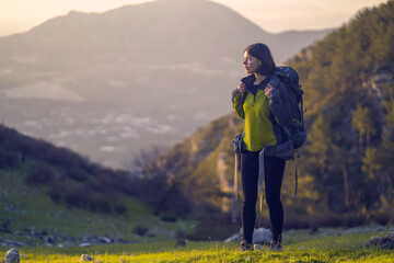 A young girl with a backpack and travel equipment looks at the amazing sunset in the mountains while she hikes along the trail. A traveler walks through the national park.