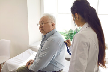 Female doctor examining elderly male patient by stethoscope at hospital ward. Physician listening breath conducting lung test. Coronavirus flu medical checkup, old people medical health insurance
