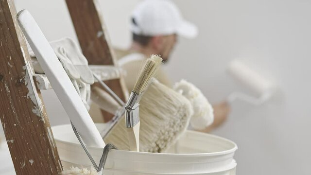 hands of house painter man decorator work of home to renovate, using roller paint and holding white bucket, a wooden ladder with paint brushes as background, close-up