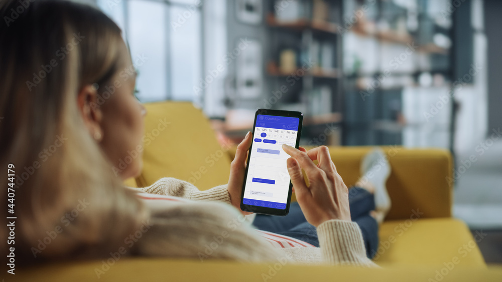 Poster Beautiful Authentic Female Using a Smartphone with Calendar Application at Home Living Room while Lying on a Couch Sofa. She's Making Plans and Checking Event Schedule for a Day.