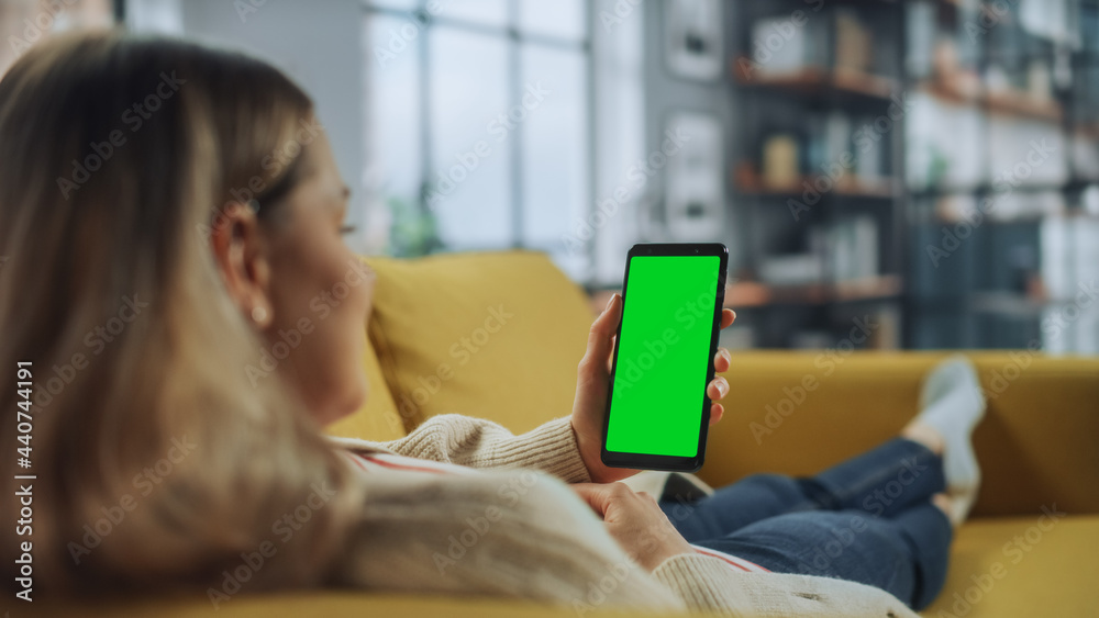 Aufkleber Beautiful Authentic Female Using a Smartphone with Green Screen Mock Up Display at Home Living Room while Lying on a Couch Sofa. She's Browsing the Internet and Checking Videos on Social Networks.