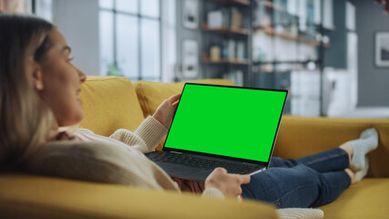 Beautiful Caucasian Specialist Working on Laptop with Green Screen Mock Up Display at Home Living Room while Lying on a Couch Sofa. Freelance Female Chatting Over the Internet on Social Networks.