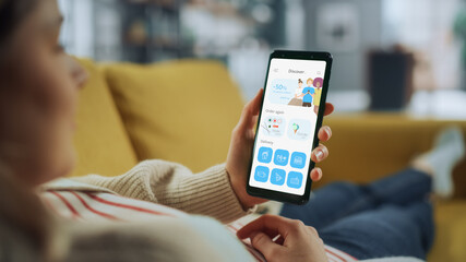 Beautiful Authentic Female Using a Smartphone with Food Delivery Application at Home Living Room while Lying on a Couch Sofa. She's Browsing the Catalog and Choosing, what to Order for a Dinner.