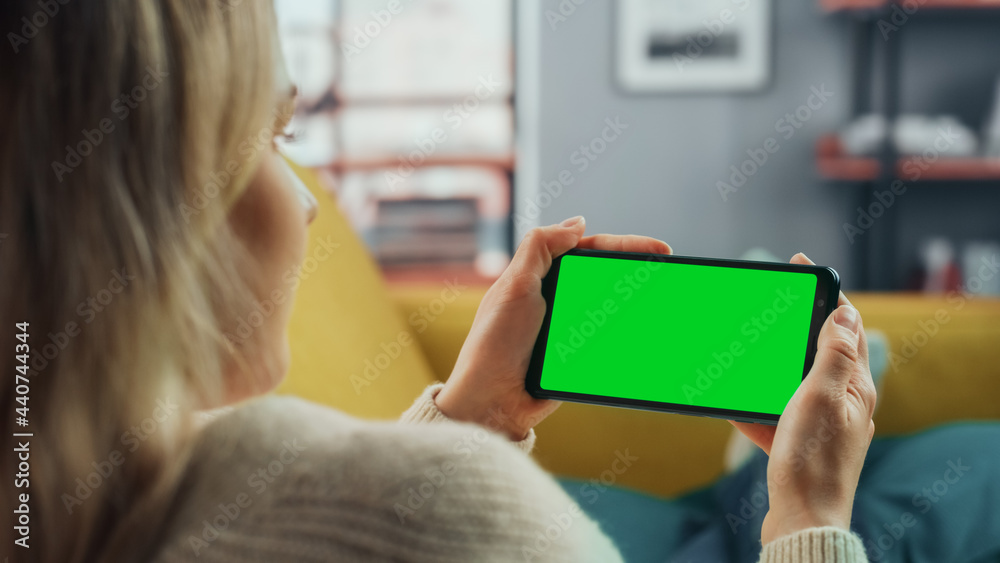 Aufkleber Beautiful Authentic Female Using a Smartphone with Green Screen Mock Up Display at Home Living Room while Lying on a Couch Sofa. She's Browsing the Internet and Checking Videos on Social Networks.