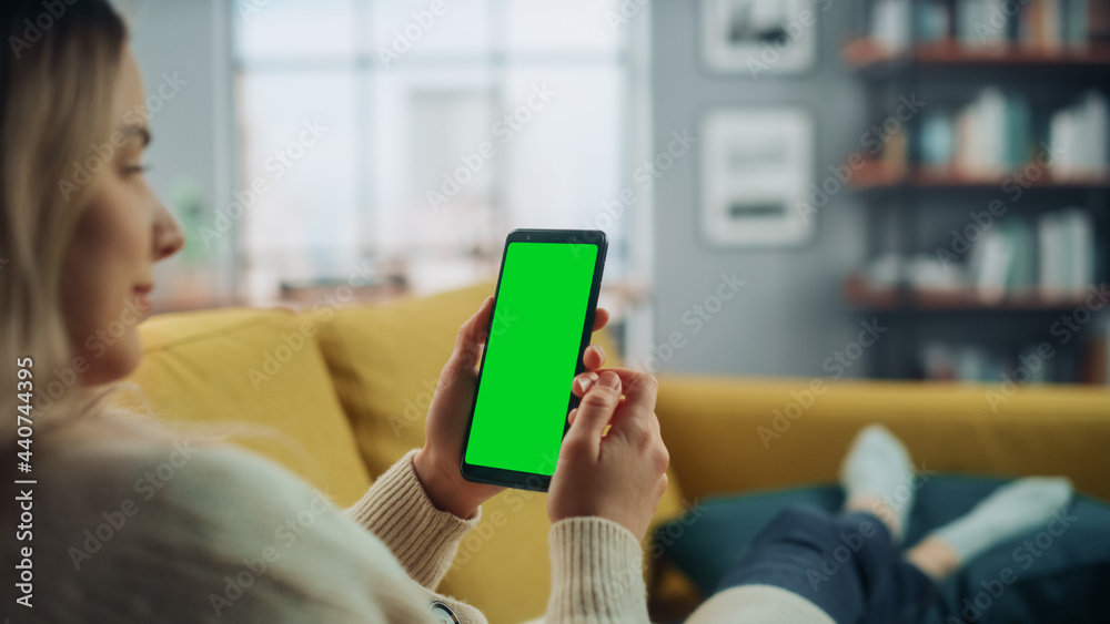 Poster Beautiful Authentic Female Using a Smartphone with Green Screen Mock Up Display at Home Living Room while Lying on a Couch Sofa. She's Browsing the Internet and Checking Videos on Social Networks.