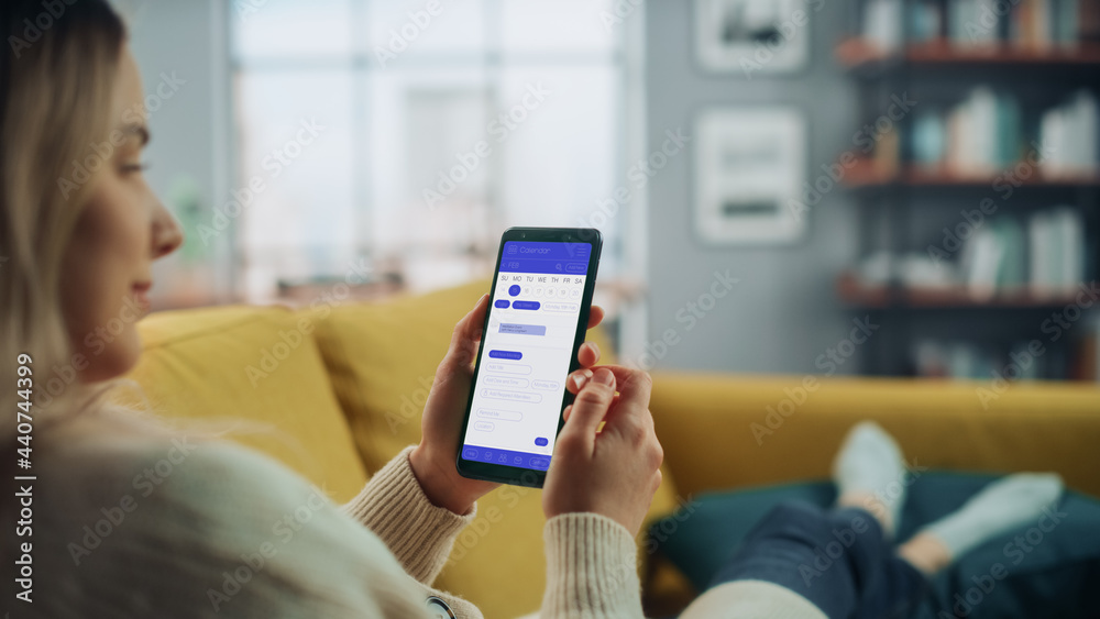 Aufkleber Beautiful Authentic Female Using a Smartphone with Calendar Application at Home Living Room while Lying on a Couch Sofa. She's Making Plans and Checking Event Schedule for a Day.