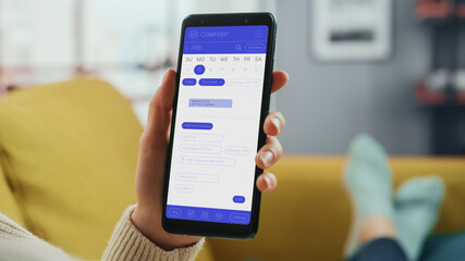 Beautiful Authentic Female Using a Smartphone with Calendar Application at Home Living Room while Lying on a Couch Sofa. She's Making Plans and Checking Event Schedule for a Day.