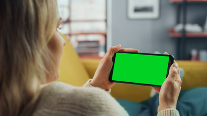 Beautiful Authentic Female Using a Smartphone with Green Screen Mock Up Display at Home Living Room while Lying on a Couch Sofa. She's Browsing the Internet and Checking Videos on Social Networks.