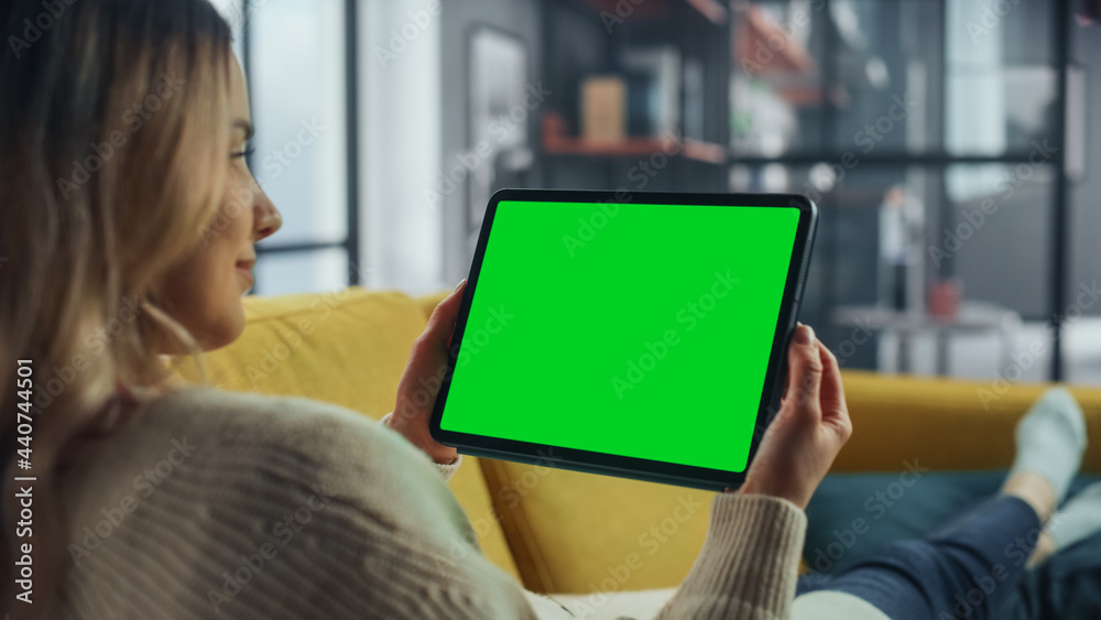 Aufkleber Beautiful Authentic Female Using Tablet Computer with Green Screen Mock Up Display at Home Living Room while Lying on a Couch Sofa. She's Browsing the Internet and Checking Videos on Social Networks.