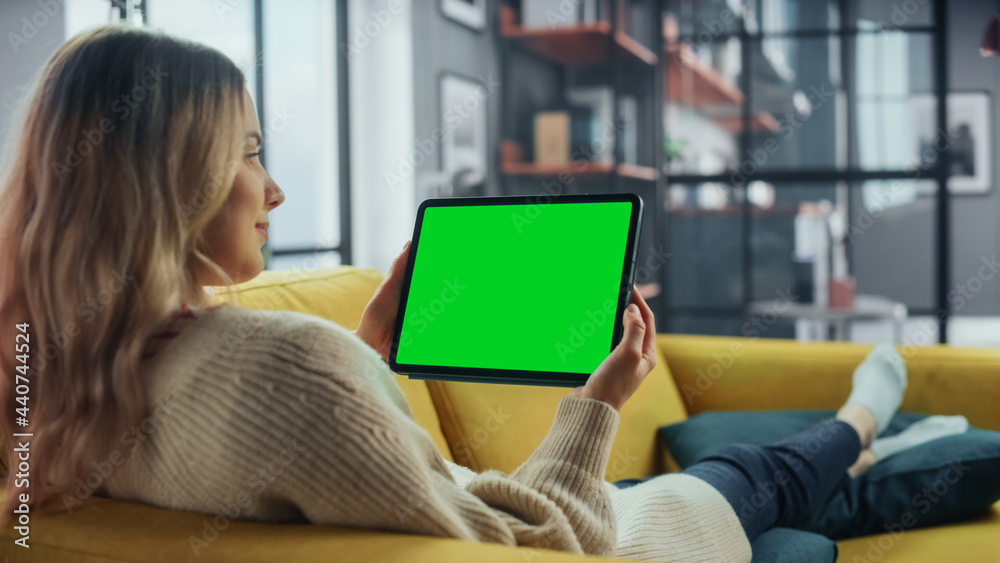 Aufkleber Beautiful Authentic Female Using Tablet Computer with Green Screen Mock Up Display at Home Living Room while Lying on a Couch Sofa. She's Browsing the Internet and Checking Videos on Social Networks.