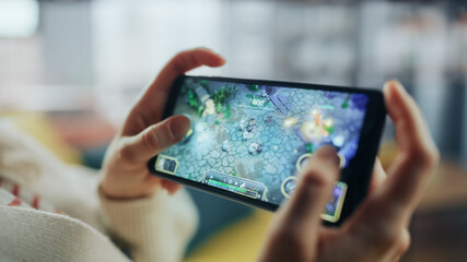 Close Up on Hands Playing an RPG Strategy Video Game on a Horizontally Held Smartphone at Home Living Room. Feminine Hands Tapping the Mobile Phone Screen with a Colorful Game Over the Internet.