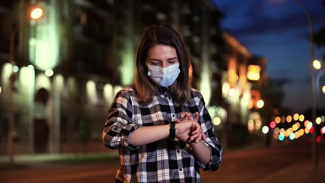 A young white-skinned girl in a protective mask stands on a city street at night and uses an electronic clock, then takes off her protective mask. Young white-skinned girl in a protective mask takes 