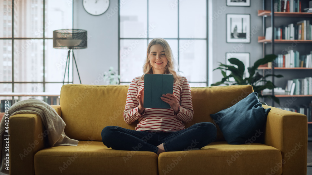 Poster beautiful caucasian female using tablet computer in stylish living room while sitting on a cozy couc
