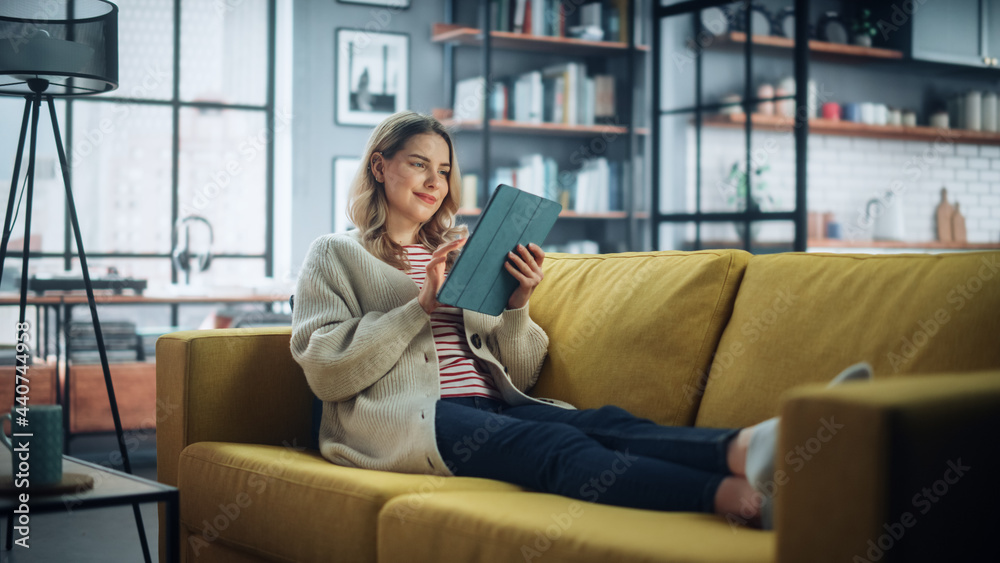 Poster beautiful caucasian female using tablet computer in stylish living room while resting on a cozy couc