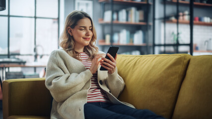 Beautiful Caucasian Female Using Smartphone in Stylish Living Room while Resting on a Cozy Couch...