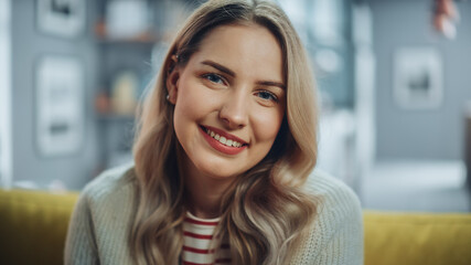 Portrait of Beautiful Young Woman with Light Brown Hair Wearing White Woolen Sweater Looking Up to the Camera and Smiling Charmingly. Successful Woman Resting in Bright Living Room.
