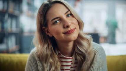 Portrait of Beautiful Young Woman with Light Brown Hair Wearing White Woolen Sweater Looking Up to the Camera and Smiling Charmingly. Successful Woman Resting in Bright Living Room.