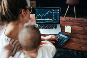 Pretty young mother with cute little baby investing in world stock market, using her laptop and...