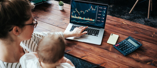Fototapeta na wymiar Pretty young mother with cute little baby investing in world stock market, using her laptop and online trading soft from home