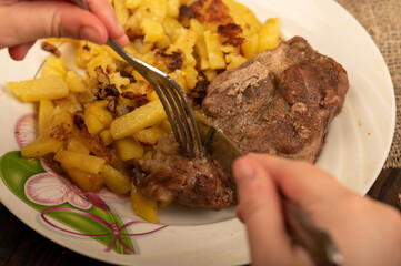 Someone is cutting a piece of juicy roast meat on a plate of fried potatoes with a knife and fork. Close-up Selective focus.