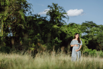 Fototapeta na wymiar Asian young pregnant woman in blue dress relaxing and enjoying life in grass nature at summer day. Healthy pregnant. Copy space