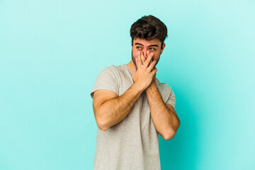 Young caucasian man isolated on blue background laughing about something, covering mouth with hands.