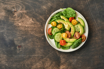 Avocado, red, yellow, black cherry tomato, spinach and cucumber fresh salad with spices pepper and olive oil in grey bowl old wooden table background. Healthy food concept. Top view.