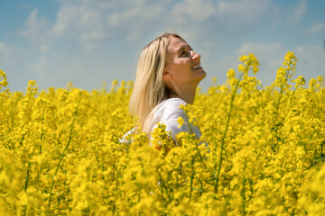 Naklejka na ściany i meble Happy smiling young blonde woman in a white shirt in a rapeseed field with her arms spread out to the side on a sunny day