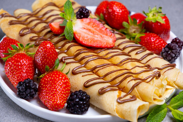 A plate of delicious pancakes or crepes rolls with fresh berries, strawberries, and chocolate on a concrete table. Top view, flat lay.