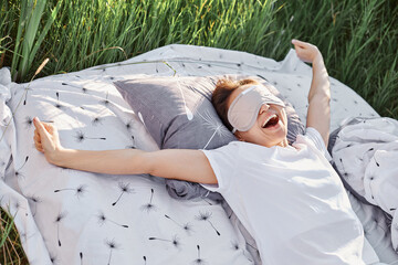 Happy yawning female wearing sleeping fold and white casual t-shirt stretching arms when waking up...