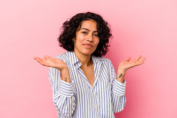 Young curly latin woman isolated on pink background confused and doubtful shrugging shoulders to...