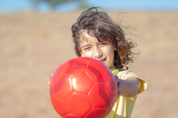 Naklejka premium little boy with long hair and yellow t-shirt plays with his red ball in a field on a sunny summer day