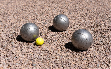 Three petanque balls (boules) close to a yellow jack target ball on a pink gravel petanque ground.