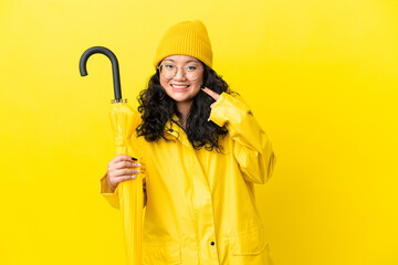 Asian woman with rainproof coat and umbrella isolated on yellow background giving a thumbs up...