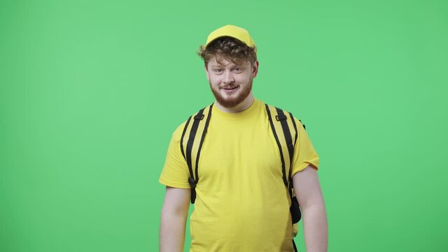 Portrait of young men waving hand and showing gesture come here. Redhead male in yellow delivery courier uniform with thermo bag posing on green screen in studio. Close up. Slow motion ready 59.97fps.