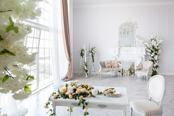 luxurious bright spacious guest room with beautiful chic furniture a huge floor-to-ceiling window in a royal style is decorated with green plants, white walls with stucco and a fireplace