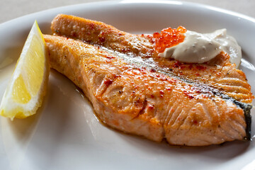 A glass of red wine and grilled salmon with white sauce, red caviar and lemon on a white plate