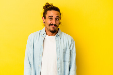 Obraz premium Young caucasian man with long hair isolated on yellow background shrugs shoulders and open eyes confused.