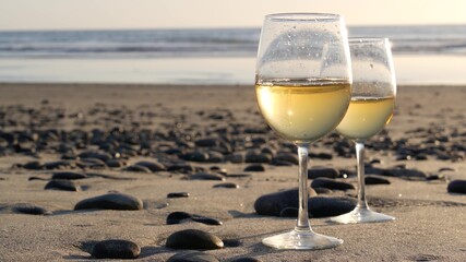 Two wineglasses on sandy ocean beach. Couple of glasses and pebbles, white wine for romantic date near sea water. Cozy beachfront sunset near tide waves. Seamless looped cinemagraph. California, USA.