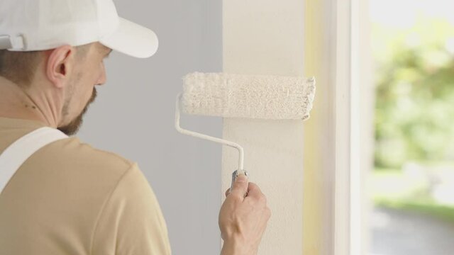 painter man decorator work of home to renovate, painting with roller paint on wall, window with green as background, close up