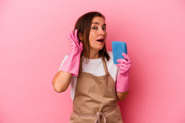 Middle age caucasian woman cleaning home isolated on pink background trying to listening a gossip.