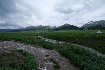 stream, green grassland and snow mountains under cloudy sky. At Nalati prairie in Xinjiang China 
