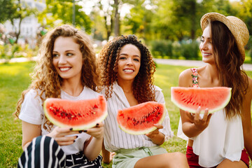 Three young woman  in the park sitting on the grass in joyful in sunny day and eating watermelon. Female friends relaxing and enjoying holidays together. People, lifestyle, travel, nature and vacation