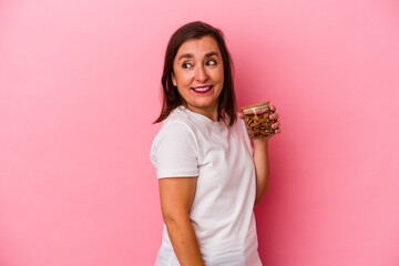 Middle age caucasian woman holding an almond jar isolated on pink background looks aside smiling, cheerful and pleasant.