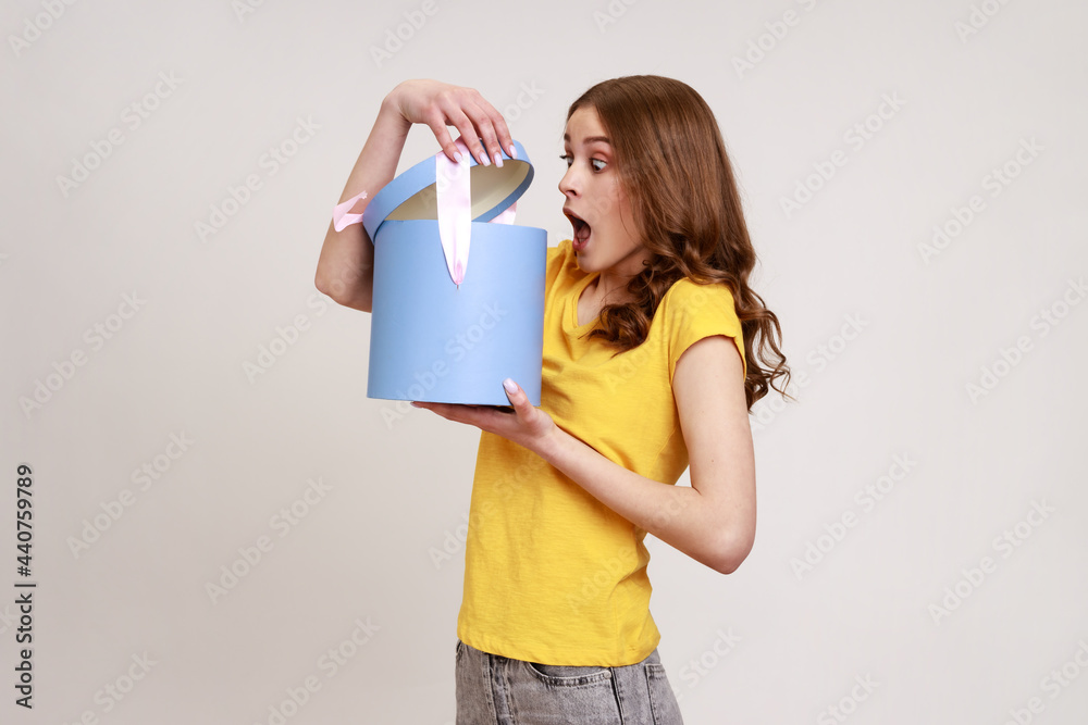 Poster Profile of shocked teenager girl in yellow casual T-shirt unpacking present box, looking inside with big eyes and open mouth, very unexpected gift. Indoor studio shot isolated on gray background.